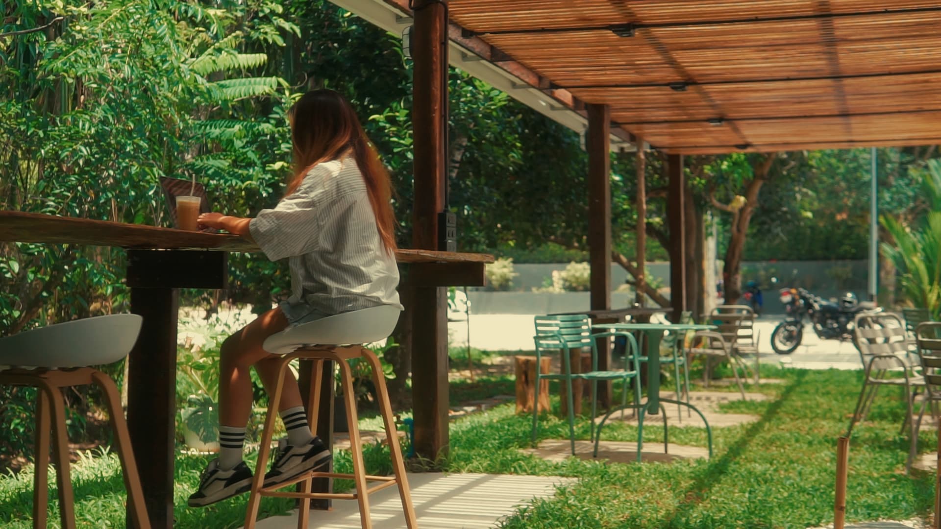 Woman working on a laptop at a wooden bar in a lush, sunlit outdoor cafe.