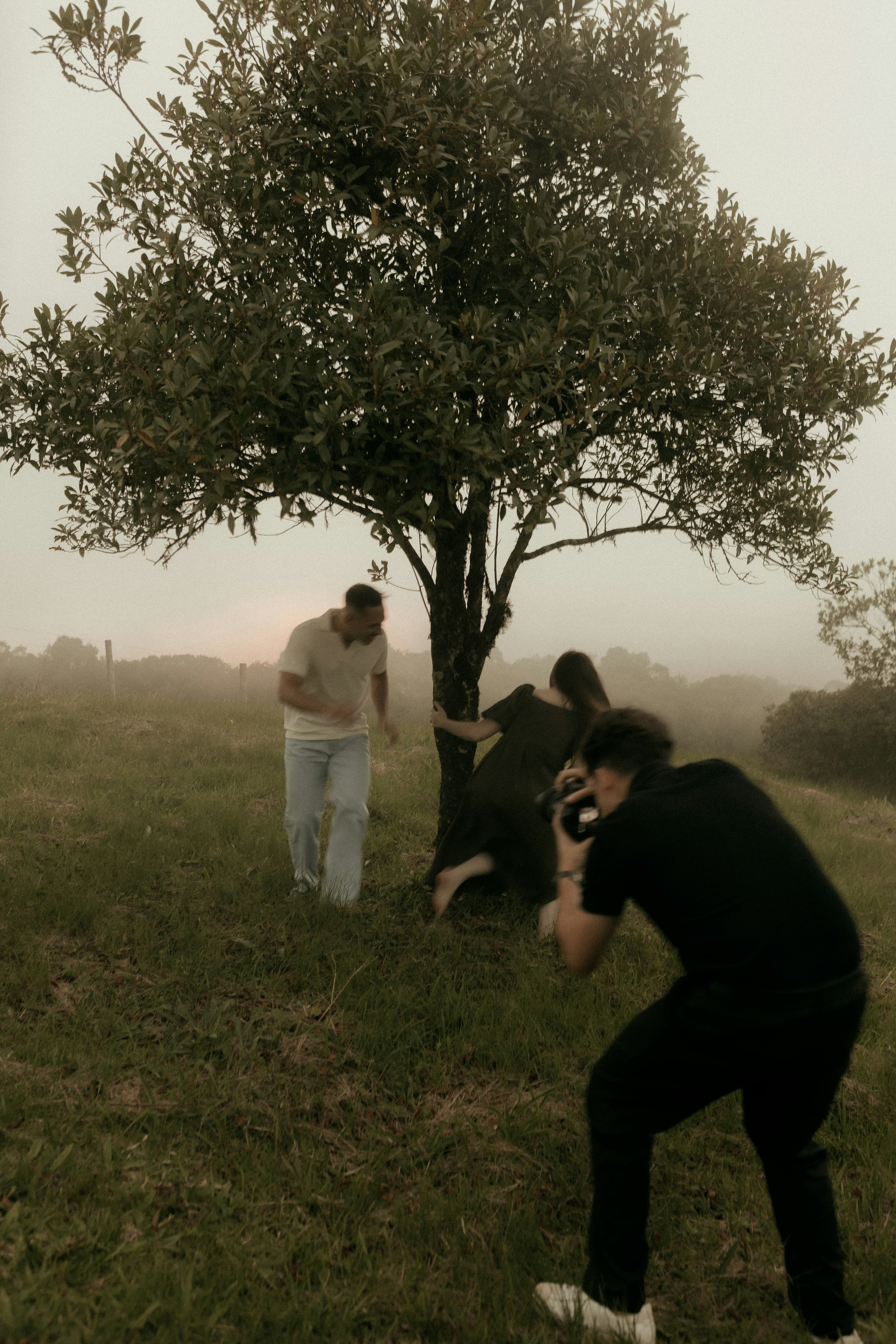Photographer captures a couple playing around a lone tree in a misty, grassy field.