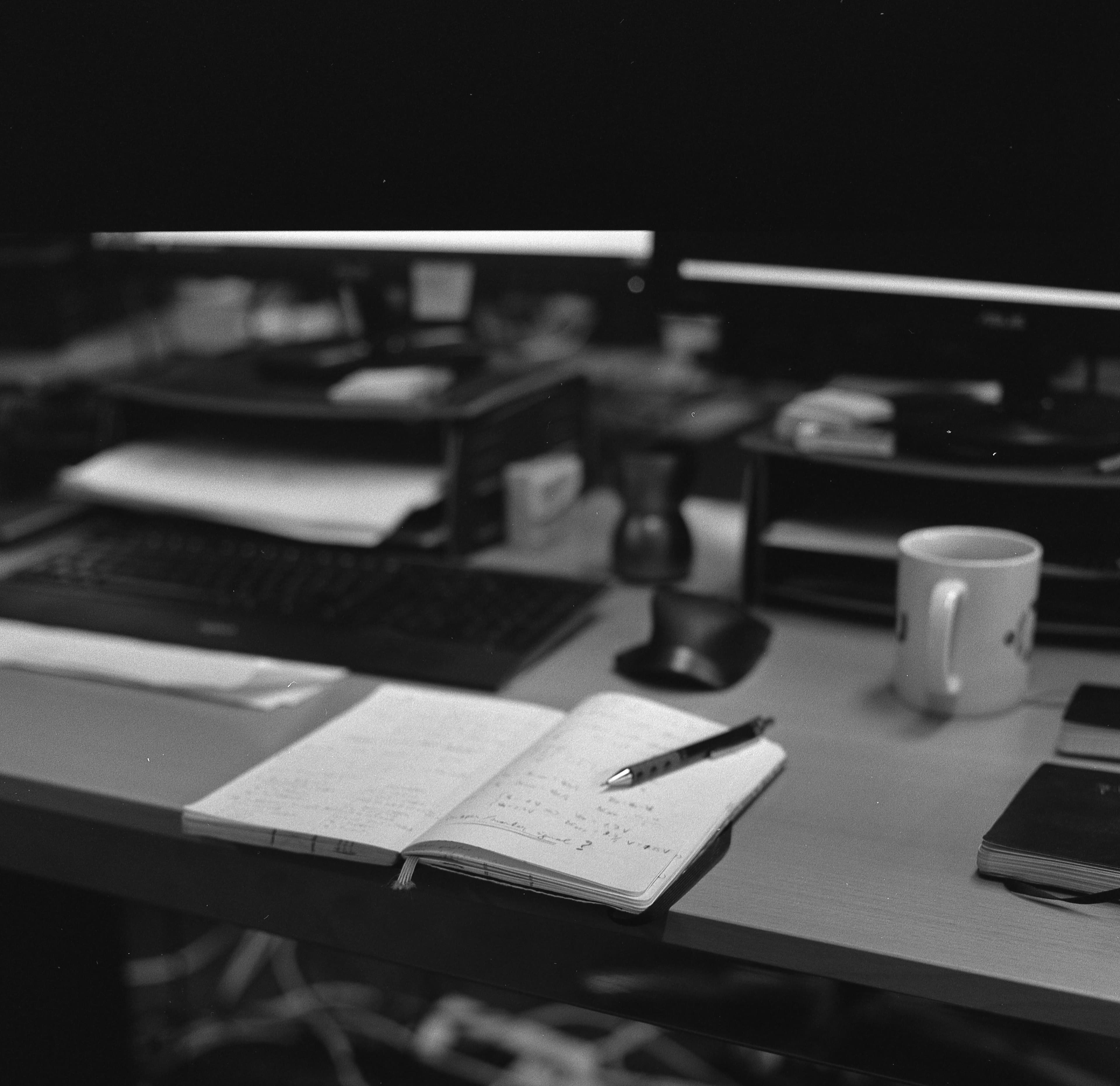 Black and white desk setup featuring an open notebook, pen, mug, and computer monitors.